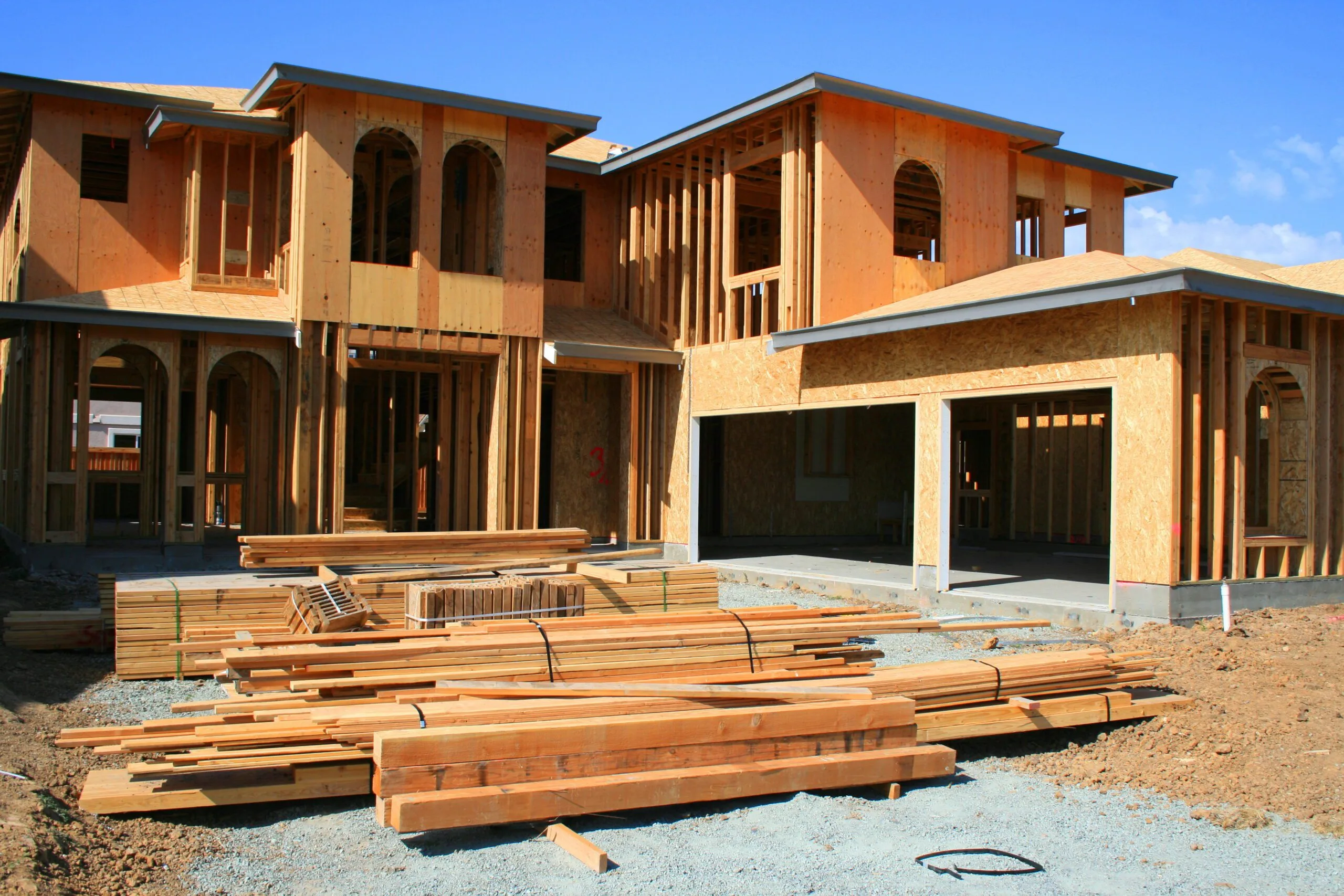 Residential construction site with partially built wooden houses and piles of lumber in the foreground.