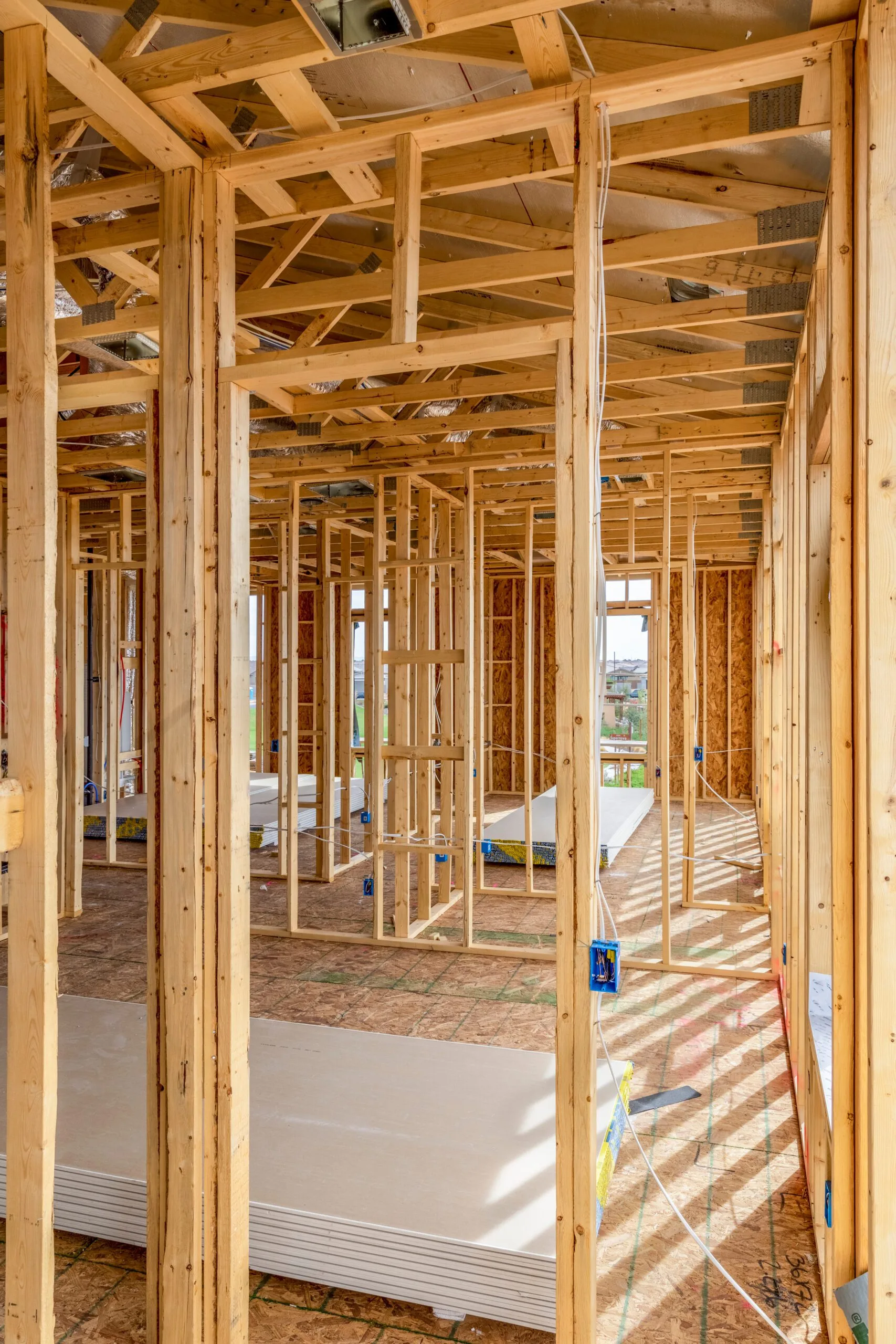 Interior view of a house under construction showing wooden frames and beams with exposed wiring and unfinished flooring.