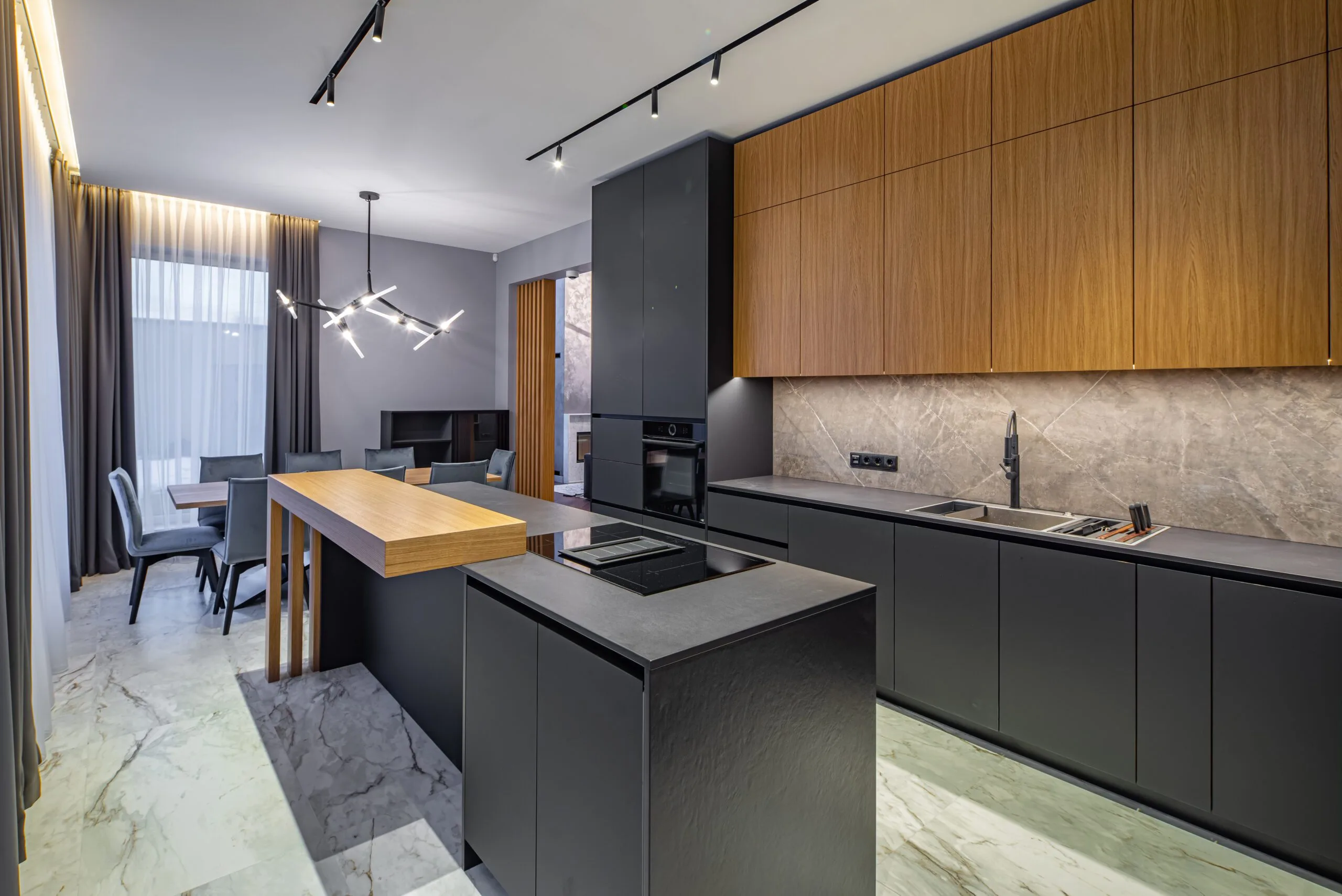 Modern kitchen with gray and wooden cabinetry, marble floors, an island, and integrated appliances, leading to a small dining area.